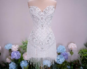 Può includere: Un abito corto bianco senza spalline con intricati decori di paillettes e perline argentate in un motivo a reticolo, scollo a cuore e orlo con lunghe frange. Una corona color oro è posizionata sopra il manichino che espone l'abito.