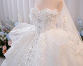 Può includere: Un abito da ballo bianco senza spalline con scollo a cuore, intricati ricami di paillettes argentate e balze in tulle a strati. L'abito è completato da un delicato girocollo di perle, un velo trasparente e un copricapo a corona dorato.