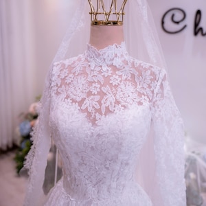May include: A white long-sleeved wedding dress featuring an intricate floral lace overlay, a high neckline, and a fitted bodice. A sheer white veil is draped over a gold-toned crown-shaped display stand positioned at the top of the dress form.