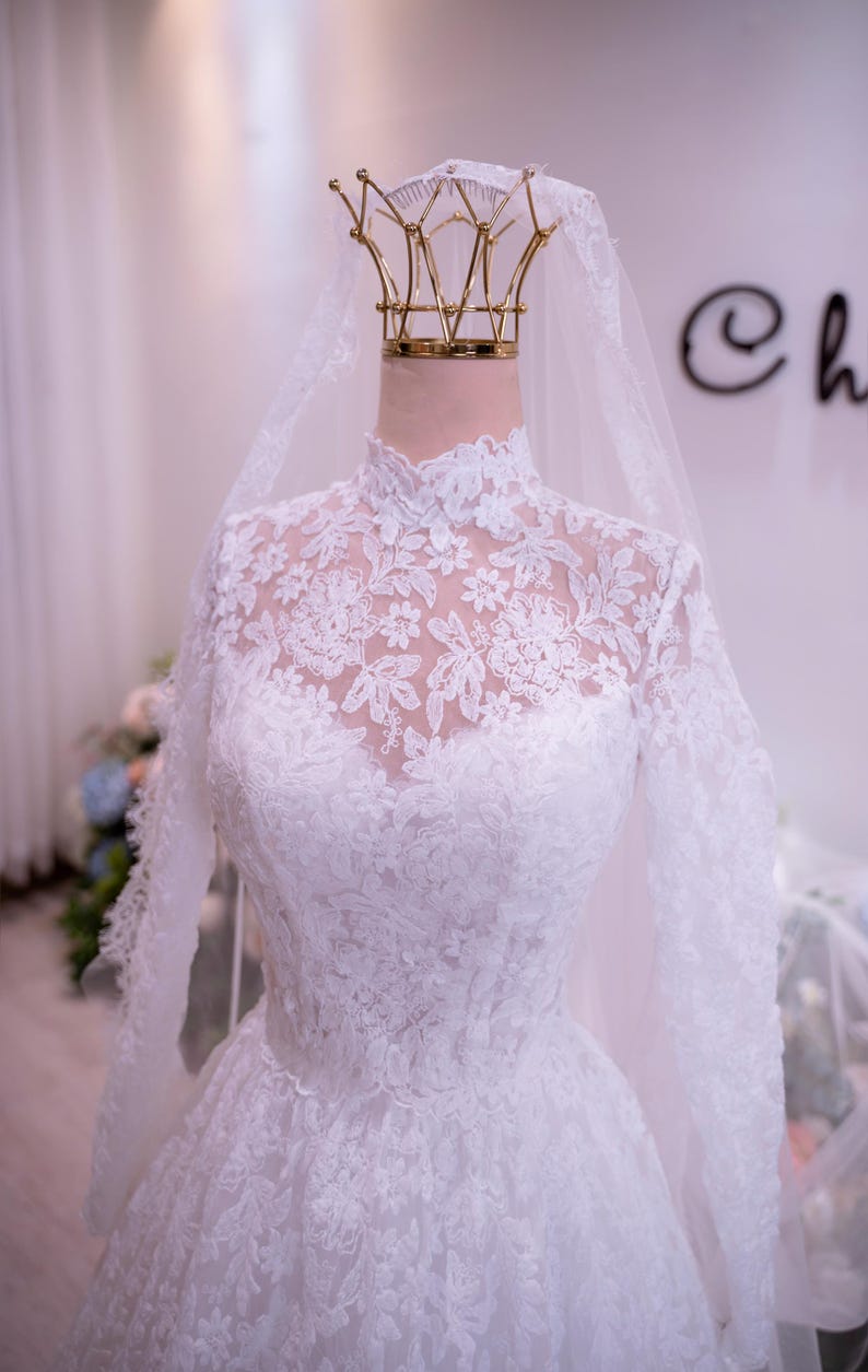 May include: A white long-sleeved wedding dress featuring an intricate floral lace overlay, a high neckline, and a fitted bodice. A sheer white veil is draped over a gold-toned crown-shaped display stand positioned at the top of the dress form.