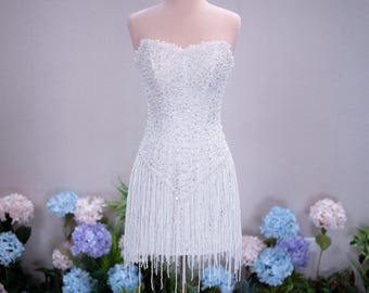Può includere: Un abito corto bianco senza spalline con scollo a cuore, interamente decorato con perline e paillettes trasparenti, e una gonna arricchita da lunghe frange bianche pendenti. Una corona in filo dorato poggia sopra l'abito su un manichino.