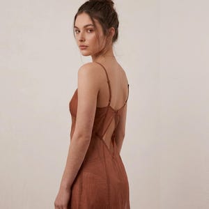 Op de afbeelding: Een roestkleurige slipdress met spaghettibandjes en een lage rug. De jurk heeft een transparante, vloeiende stof en een getailleerd lijfje. Het model kijkt in de camera, met haar haar in een knot.