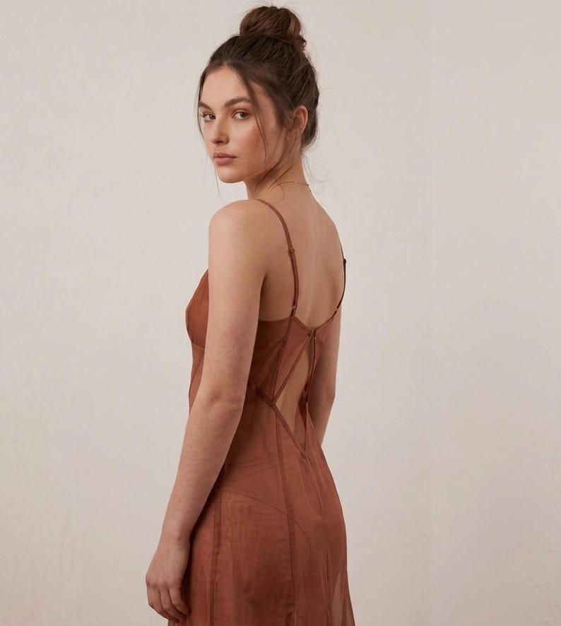 Op de afbeelding: Een roestkleurige slipdress met spaghettibandjes en een lage rug. De jurk heeft een transparante, vloeiende stof en een getailleerd lijfje. Het model kijkt in de camera, met haar haar in een knot.