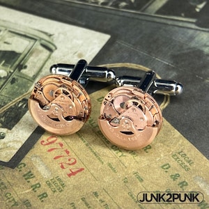 May include: A pair of cufflinks featuring circular rose gold watch movements with visible gears and internal components. Each piece is mounted on a polished silver-tone metal toggle backing. The watch faces display the text SEVENTEEN JEWELS.