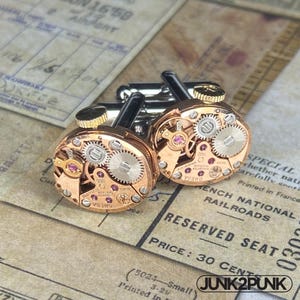 May include: A pair of cufflinks featuring the intricate internal mechanisms of a watch. The gold-toned metal components, including gears and jewels, are visible. The cufflinks have a silver backing and are set against a vintage paper background.