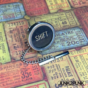 May include: A silver tie clip with a black, circular button displaying the word "SHIFT" in gold. The clip includes a chain and a rectangular bar. The background is made up of vintage ticket stubs.