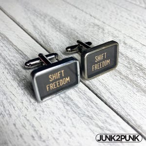 May include: A pair of cufflinks with a rectangular black face and silver-tone metal trim. The text "SHIFT FREEDOM" is printed in gold on each cufflink. The cufflinks are displayed on a light grey wooden surface.
