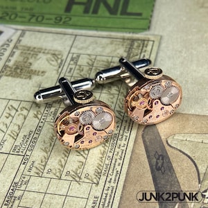 May include: A pair of cufflinks crafted from vintage rose gold-toned watch movements. Each oval-shaped piece features exposed silver and gold gears, small purple jewels, and a winding crown with an Omega logo. The silver-toned T-bar fasteners are attached to the back of the watch mechanisms.