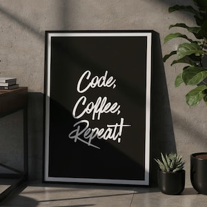 Può includere: Un poster nero incorniciato con la scritta "Code, Coffee, Repeat!" è esposto accanto a una scrivania con un laptop e cuffie. Una pianta in vaso e una piccola succulenta sono anche nell'immagine, creando una scena di lavoro.