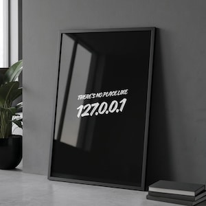 Könnte beinhalten: Ein schwarzes Poster mit weißem Text "THERE'S NO PLACE LIKE 127.0.0.1" lehnt an einer grauen Wand. Das Poster befindet sich in einem modernen Büroumfeld mit Schreibtisch, Laptop und Topfpflanze.
