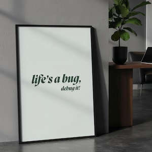 May include: A framed print with the text "life's a bug, debug it!" in a dark teal font. The print is in a black frame and leans against a grey wall. The background includes a desk with a laptop and a potted plant.