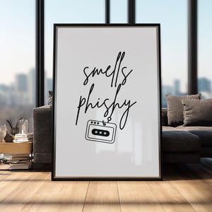 May include: A framed art print with the words "smells phishy" in black script. Below the text is a black and white illustration of a cassette tape. The print is displayed in a living room setting with a window and a couch.