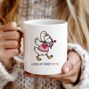 May include: White ceramic mug featuring a cartoon duck dressed in a pink sweater and heart-shaped chest, holding a bow and arrow. The text "LOVE AT FIRST BYTE" is printed below the duck. The mug is held by a person.