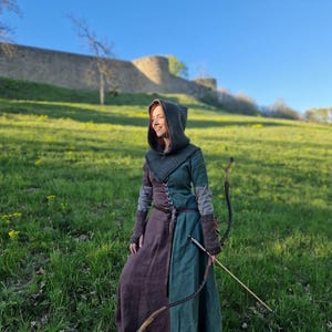 May include: A medieval-style dress with a hooded cloak, brown and green color-blocked design, and a brown belt. The person is holding a bow and arrow, standing in a grassy field with a stone wall in the background.
