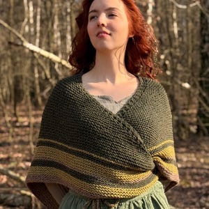 May include: A knitted wrap in olive green and gold. The wrap has horizontal stripes and a V-neck design. The garment is worn over a green dress, creating a layered look. The background is a blurred forest scene.