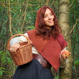 May include: A woman in a forest setting, wearing a rust-colored hooded shawl, a dark skirt, and a brown leather belt. She holds a woven basket and an apple. The scene evokes a fairytale or historical theme.