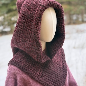 May include: A burgundy knit hood and cowl set. The hood features a textured stitch and a deep cowl that drapes over the shoulders. The set is displayed on a mannequin, with a blurred outdoor background.