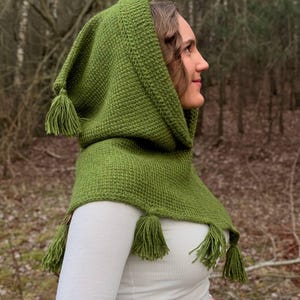 May include: A green knitted hood with tassels. The hood is a shade of olive green and has a textured pattern. The tassels are at the end of the hood and on the shoulders. The hood is worn over a white long-sleeved shirt.