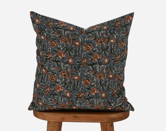 May include: Square throw pillow with a floral pattern in shades of gray, rust, and cream. The pillow is resting on a small, round wooden stool. The floral design features stylized flowers and leaves.