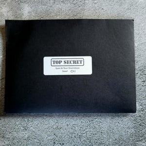 Puede incluir: Un sobre negro con una etiqueta blanca que dice "TOP SECRET". La etiqueta también incluye el texto "Open At Your Discretion" y "Case # 011". El sobre está sobre una superficie gris.