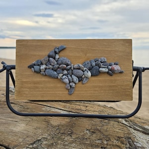 Peut inclure: Plaque en bois ornée d'une mosaïque de pierres grises et brunes représentant la péninsule supérieure du Michigan. Montée sur un support en métal noir, elle se détache sur une plage.