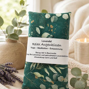 Könnte beinhalten: Ein rechteckiges Augenkissen aus dunkelgrünem Stoff mit einem Muster aus Eukalyptusblättern und goldenen Akzenten. Ein weißes Papierband umschließt die Mitte mit dem Text: Lavendel Relax Augenkissen, Yoga - Meditation - Entspannung, Bezug: 100 % Baumwolle, Füllung: Bio-Leinsamen und getrockneten Lavendelblüten, Anwendung warm und kalt.