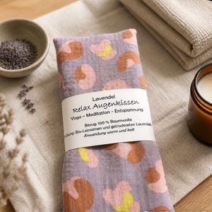 Könnte beinhalten: Ein rechteckiges Augenkissen mit lavendelfarbenem Hintergrund und einem Muster aus braunen, rosafarbenen und gelben abstrakten Formen. Ein weißes Papieretikett ist mittig umwickelt mit dem Text: Lavendel Relax Augenkissen Yoga - Meditation - Entspannung.