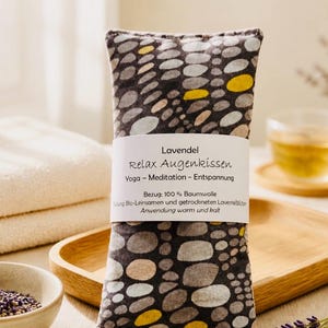 Könnte beinhalten: Ein rechteckiges Augenkissen mit einem Kieselsteinmuster in Grau, Gelb und Braun. Das Kissen hat ein weißes Etikett mit dem Text "Lavendel Relax Augenkissen" und weiteren deutschen Texten. Es ist für Yoga, Meditation und Entspannung gedacht.