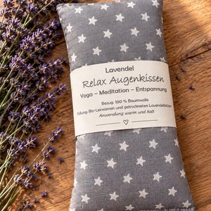 Könnte beinhalten: Ein rechteckiges graues Augenkissen mit weißem Sternenmuster, umwickelt mit einer Papierbanderole mit der Aufschrift Lavendel Relax Augenkissen, Yoga - Meditation - Entspannung, 100% Baumwolle, Bio-Leinsamen und getrockneten Lavendelblüten.