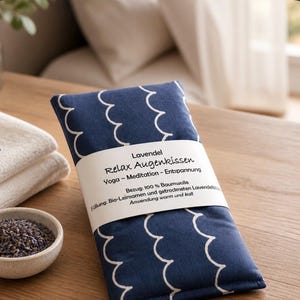 Könnte beinhalten: Ein rechteckiges, marineblaues Augenkissen mit weißem Wellenmuster. Eine weiße Papierbanderole umschließt die Mitte mit dem Text: Lavendel Relax Augenkissen, Yoga - Meditation - Entspannung, Bezug: 100 % Baumwolle, Füllung: Bio-Leinsamen und getrockneten Lavendelblüten, Anwendung warm und kalt.