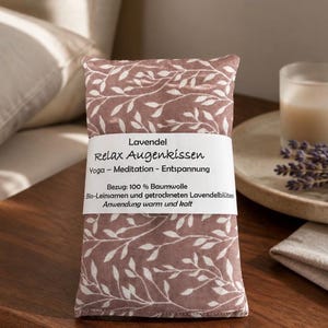 Könnte beinhalten: Ein rechteckiges Augenkissen aus altrosa Stoff mit weißem Blattmuster. Eine weiße Papierbanderole umschließt die Mitte mit dem Text: Lavendel Relax Augenkissen Yoga - Meditation - Entspannung Bezug: 100 % Baumwolle Bio-Leinsamen und getrockneten Lavendelblüten Anwendung warm und kalt.