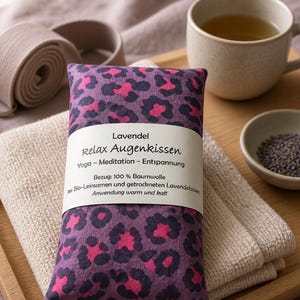 Könnte beinhalten: Ein violettes Augenkissen mit pinkfarbenem Leopardenmuster, beschriftet mit "Lavendel Relax Augenkissen". Das Bild zeigt eine Tasse Tee, eine kleine Schale mit Lavendelknospen, einen Yogagurt und ein gefaltetes Handtuch, alles auf einem Holztablett angeordnet.
