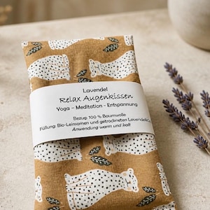 Könnte beinhalten: Ein rechteckiges Augenkissen aus senfgelbem Stoff mit weiß-schwarzem Leopardenmuster. Eine weiße Papierbanderole umschließt die Mitte mit dem Text: Lavendel Relax Augenkissen Yoga - Meditation - Entspannung.