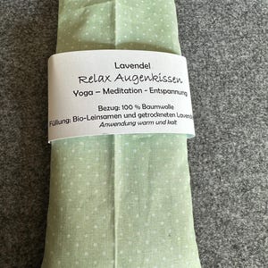 Könnte beinhalten: Ein hellgrünes Augenkissen mit weißen Punkten. Das Kissen hat ein weißes Etikett mit dem Text "Lavendel Relax Augenkissen Yoga - Meditation - Entspannung". Es besteht aus 100 % Baumwolle und ist mit Leinsamen und getrocknetem Lavendel gefüllt.
