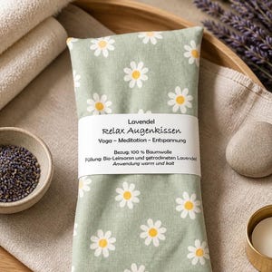 Pode incluir: Uma almofada retangular para os olhos em tecido verde sálvia com um padrão de margaridas brancas e amarelas. Uma faixa de papel branca envolve o centro com o texto: Lavendel Relax Augenkissen, Yoga - Meditation - Entspannung, Bezug: 100 % Baumwolle, Füllung: Bio-Leinsamen und getrockneten Lavendel, Anwendung warm und kalt.