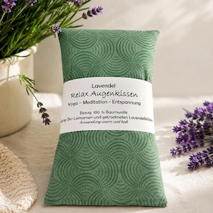 Könnte beinhalten: Ein rechteckiges grünes Augenkissen mit einem sich wiederholenden fächerförmigen Muster. Ein weißes Papierband umschließt die Mitte mit dem Text: Lavendel Relax Augenkissen, Yoga - Meditation - Entspannung, Bezug: 100 % Baumwolle, Bio-Leinsamen und getrockneten Lavendelblüten, Anwendung warm und kalt.