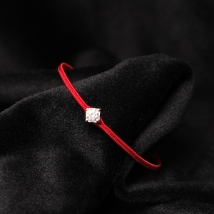 Może przedstawiać: Czerwona bransoletka z kwadratowym, przezroczystym kamieniem szlachetnym pośrodku. Bransoletka wykonana jest z cienkiego, tkanego czerwonego sznurka. Tło to ciemna, teksturowana tkanina.