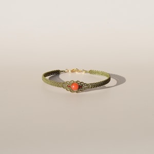 Può includere: Bracciale in macramè verde oliva fatto a mano con una perla centrale color corallo. Il bracciale presenta una chiusura color oro e dettagli decorativi attorno alla perla. Il bracciale è esposto su uno sfondo neutro.