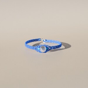 Op de afbeelding: Een blauwe macramé armband met een heldere, ronde kraal in het midden. De armband is gemaakt van geweven blauwe draden en heeft kleine turquoise kralen. De armband heeft een omtrek van ongeveer 18 cm.
