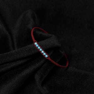 Può includere: Un braccialetto rosso intrecciato con cinque perline azzurre. Il braccialetto è posto su uno sfondo scuro e strutturato, evidenziando il contrasto tra i colori e le trame. Un gioiello semplice ma elegante.