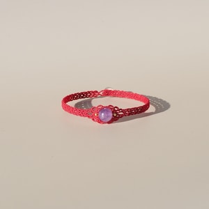 Può includere: Un braccialetto macramè rosso fatto a mano con una gemma viola centrale. Il braccialetto è finemente annodato con un motivo delicato e ha una piccola chiusura. La gemma è rotonda e incastonata in una montatura color oro.