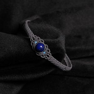 Peut inclure: Un bracelet macramé gris avec une pierre précieuse bleu foncé centrale et de petites perles turquoise. Le bracelet est posé sur un fond sombre, soulignant le nœud complexe et les couleurs vives des pierres.
