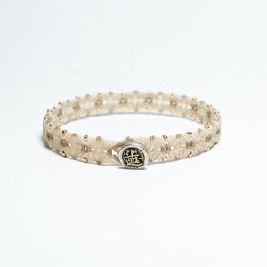 Puede incluir: Una pulsera beige con un diseño tejido, con pequeñas cuentas doradas y un cierre plateado con un diseño en relieve. La pulsera es circular y descansa sobre un fondo blanco.