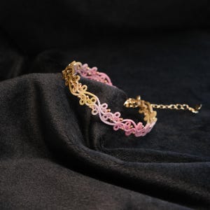 May include: A delicate bracelet with a floral design, featuring a gradient of colors from gold to pink. The bracelet is made of intricate lace-like material and has small gold beads. The background is a dark, textured fabric.