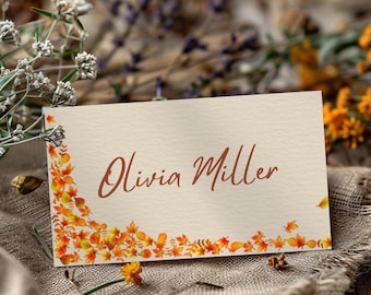 May include: A beige place card with the name "Olivia Miller" in brown script. The card is decorated with a border of autumn leaves in shades of orange, yellow, and red. The background includes flowers and a textured fabric.