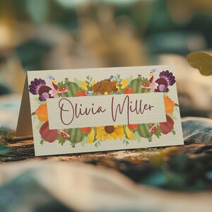May include: A folded place card with a harvest design. The card has a floral border with pumpkins, turkeys, and sunflowers. The name "Olivia Miller" is written in a cursive font across the centre.