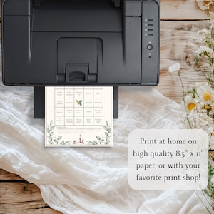 May include: A printed bingo card with a botanical design is shown emerging from a black printer. The card features a grid of squares with various words and images. Text on the card reads "Print at home on high quality 8.5" x 11" paper, or with your favorite print shop!"
