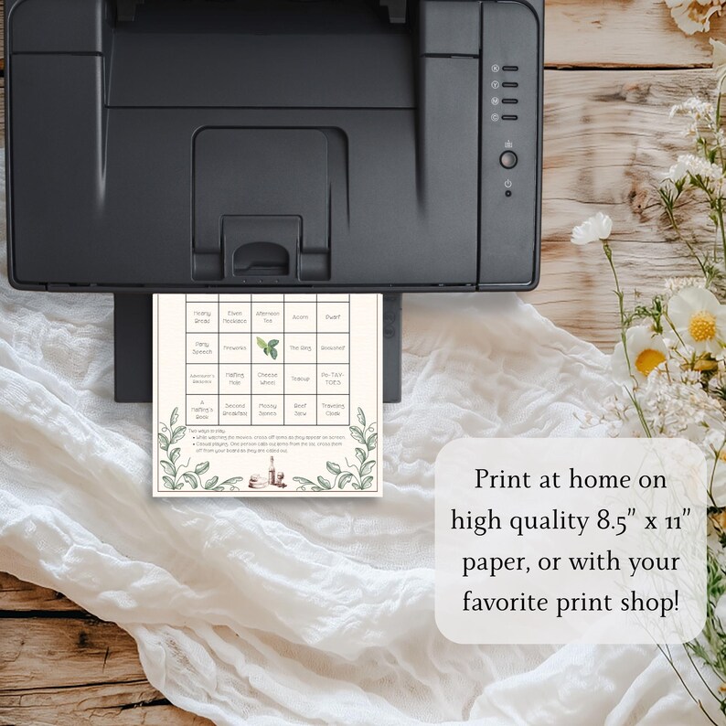 May include: A printed bingo card with a botanical design is shown emerging from a black printer. The card features a grid of squares with various words and images. Text on the card reads "Print at home on high quality 8.5" x 11" paper, or with your favorite print shop!"