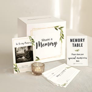 May include: A white memory box with a slot, a framed sign that says "Share a Memory", and a sign that says "Memory Table". The set includes a photo, a candle holder, and memory cards. Green leaf accents and gold trim are featured.