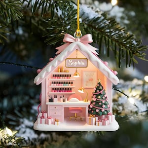 Könnte beinhalten: Ein rosafarbener, hausförmiger Weihnachtsbaumschmuck mit einem Nagelstudio-Interieur, Regalen voller Nagellack, einem Schreibtisch, einem Stuhl und einem geschmückten Weihnachtsbaum. Das Schild über dem Schreibtisch zeigt den Namen Sophia.